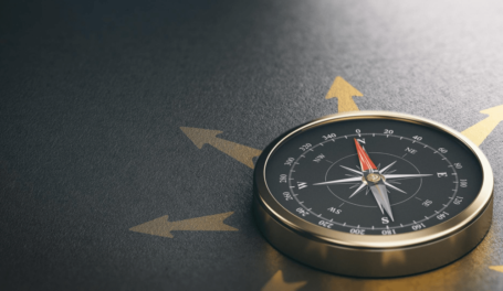 A gold-rimmed compass with a black face and red needle pointing north is placed on a dark surface with directional arrows.