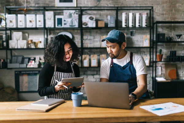 Small business owners in a coffee shop using laptop and tablet to manage orders, inventory, and digital operations together